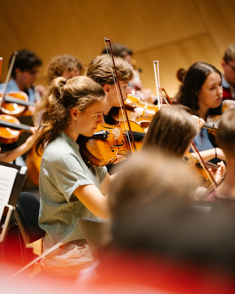 Ein Orchester (Bundesjugendorchester) spielt klassische Musik. Mehrere junge Musiker konzentrieren sich auf ihre Instrumente, vor allem Geigen, während eines Auftritts.