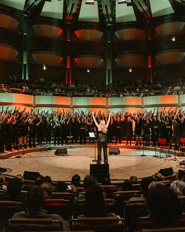 Veedelperlen in der Kölner Philharmonie: A conductor leads a large choir in a modern concert hall. The audience sits attentively, as colorful lights illuminate the scene.
