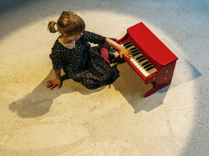 Ein kleines Kind sitzt auf dem Boden und spielt ein rotes Kinderklavier im Scheinwerferlicht.