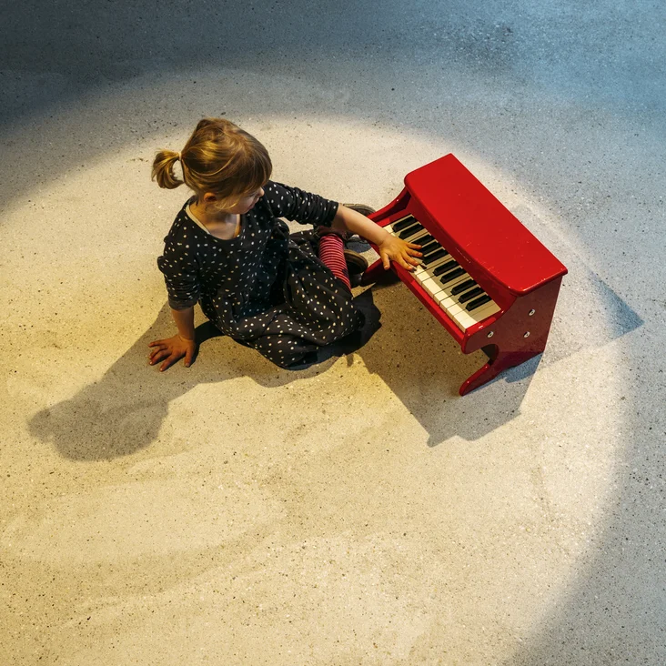 Ein kleines Kind sitzt auf dem Boden und spielt ein rotes Kinderklavier im Scheinwerferlicht.