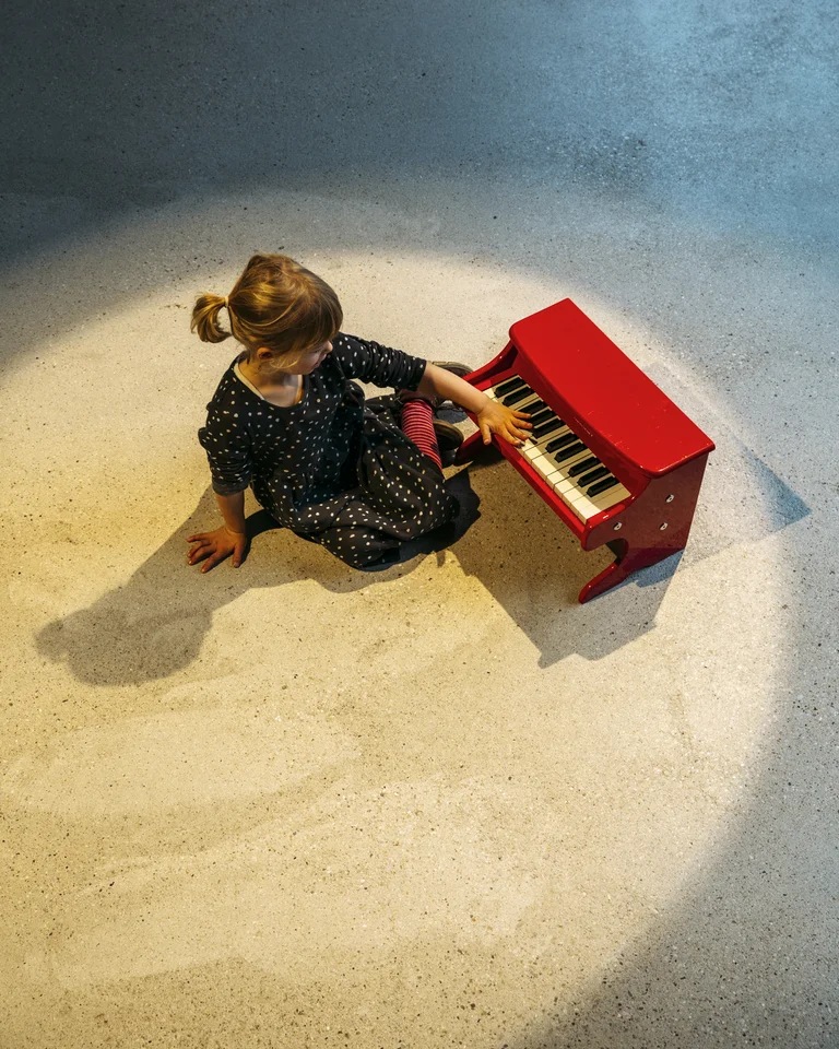 Ein kleines Kind sitzt auf dem Boden und spielt ein rotes Kinderklavier im Scheinwerferlicht.