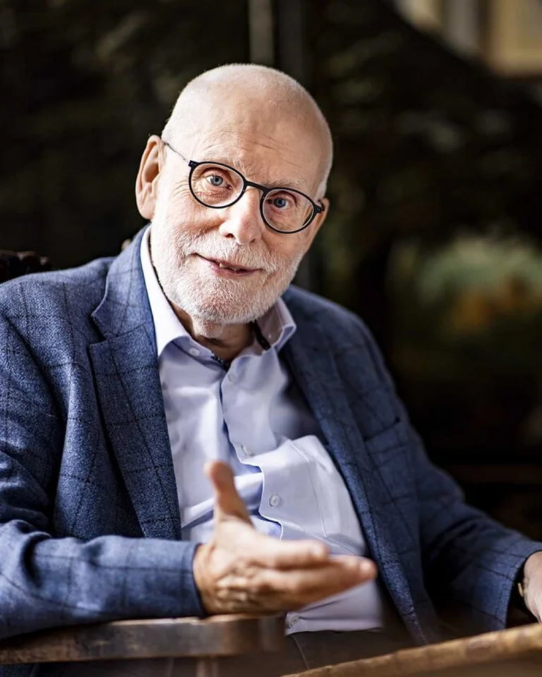 Ein älterer Mann mit Brille und Bart sitzt in einem Raum mit Kunstwerken im Hintergrund und spricht gestikulierend.