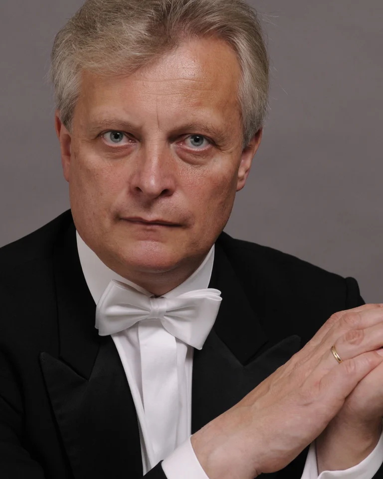 A man in a black tuxedo and bow tie sits with his hands clasped, against a gray background, gazing directly at the camera.