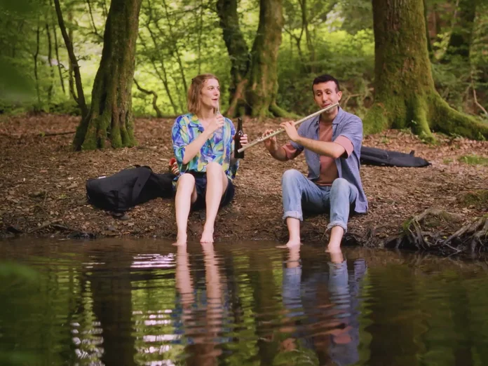 Two people sit on the bank of a tranquil forest pond, playing flutes. The wooded background enhances the peaceful atmosphere.