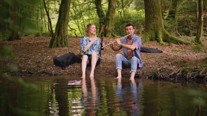 Zwei Menschen sitzen am Ufer eines ruhigen Waldsees und spielen entspannt Flöte. Der bewaldete Hintergrund verstärkt die friedliche Atmosphäre.