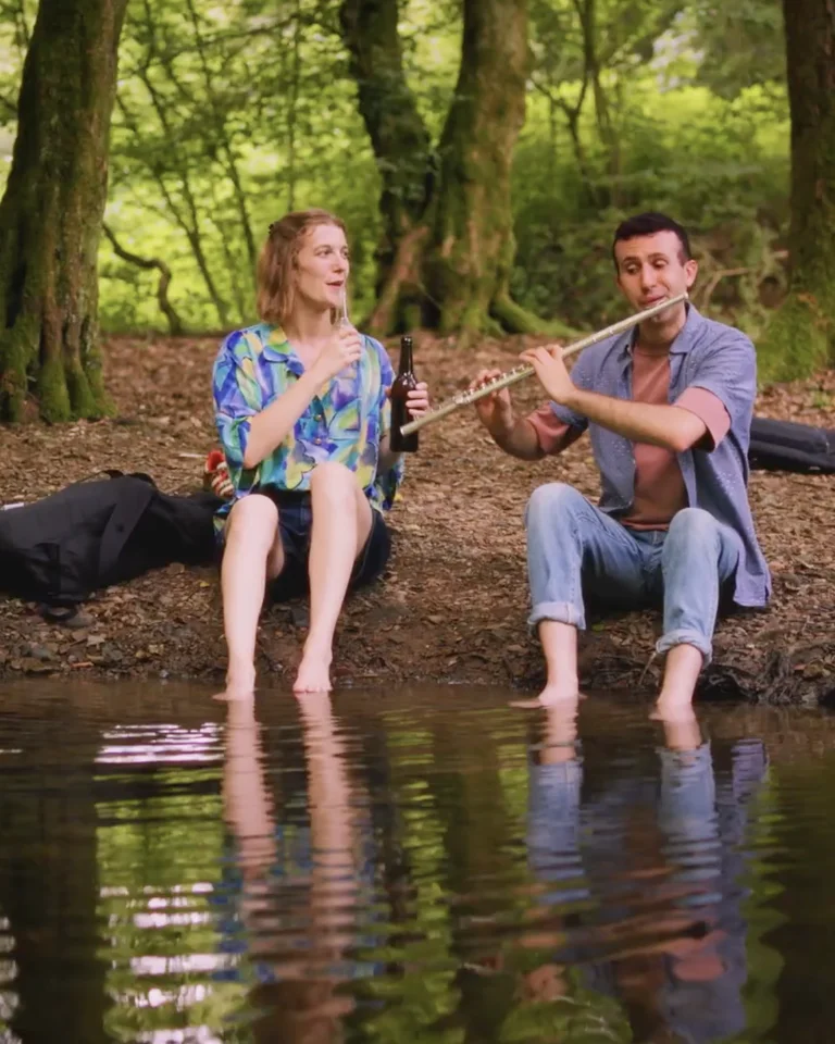 Zwei Menschen sitzen am Ufer eines ruhigen Waldsees und spielen entspannt Flöte. Der bewaldete Hintergrund verstärkt die friedliche Atmosphäre.