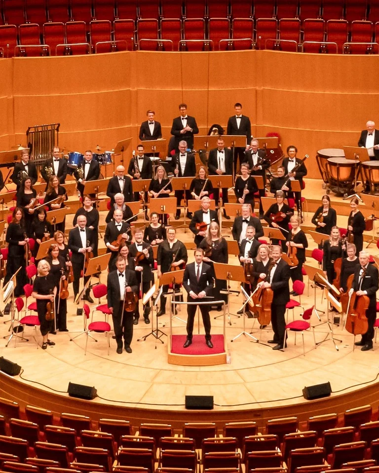 Ein Orchester (Ford-Sinfonieorchester) steht auf der Bühne eines großen Konzertsaals, bereit für eine Aufführung. Musiker tragen formelle Kleidung.