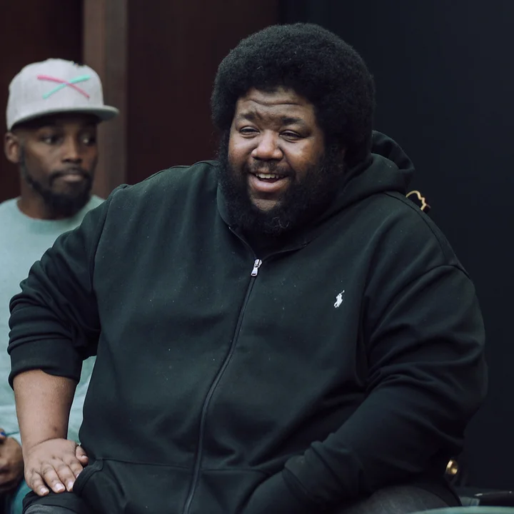 Tyshawn Sorey, wearing a black hoodie, is laughing and sitting indoors. Another person in the background is wearing a baseball cap.
