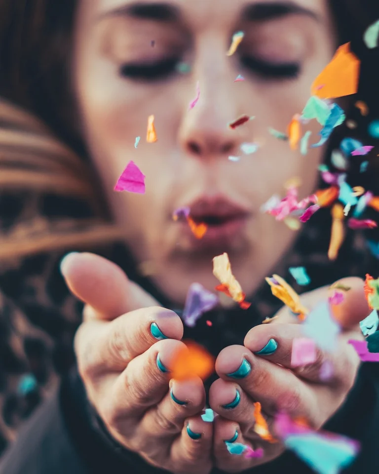 Frau pustet bunte Konfetti in die Luft. Lebhafte Farben schweben um ihr Gesicht, Ausdruck von Freude und Feierlichkeit.