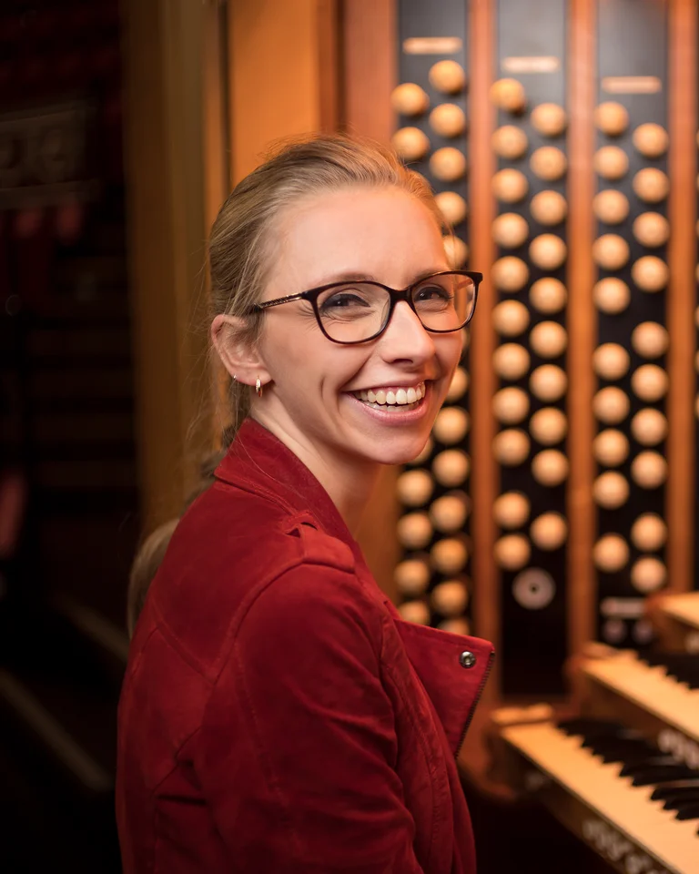 Eine lächelnde Person mit Brille sitzt an einer imposanten Orgel, während sie Partituren auf einem Bildschirm betrachtet.