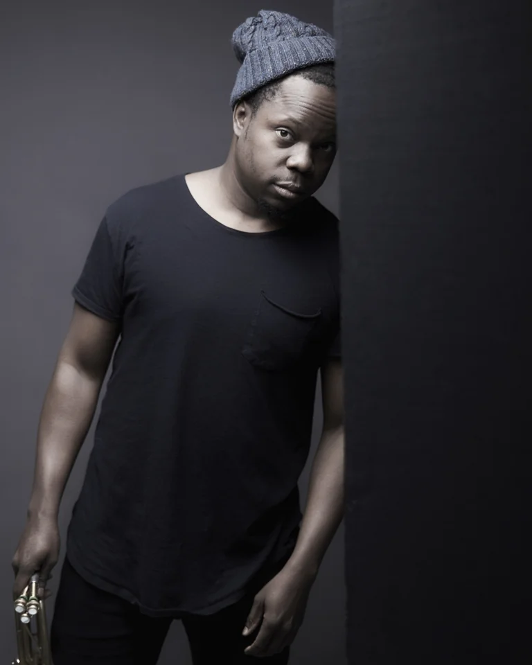 A man in a black T-shirt and gray beanie leans casually against a wall holding a trumpet.