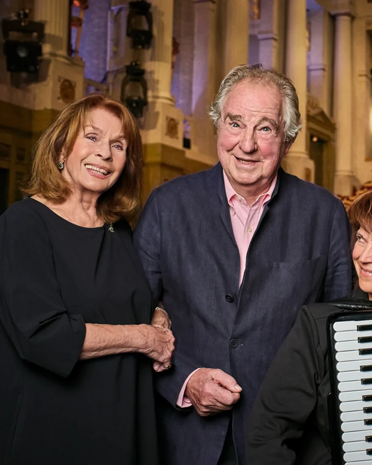 Drei Personen (Senta Berger | Friedrich von Thun | Maria Reiter) stehen vor einer Bühne in einem Theater. Eine Person hält ein Akkordeon, während die anderen lächeln.