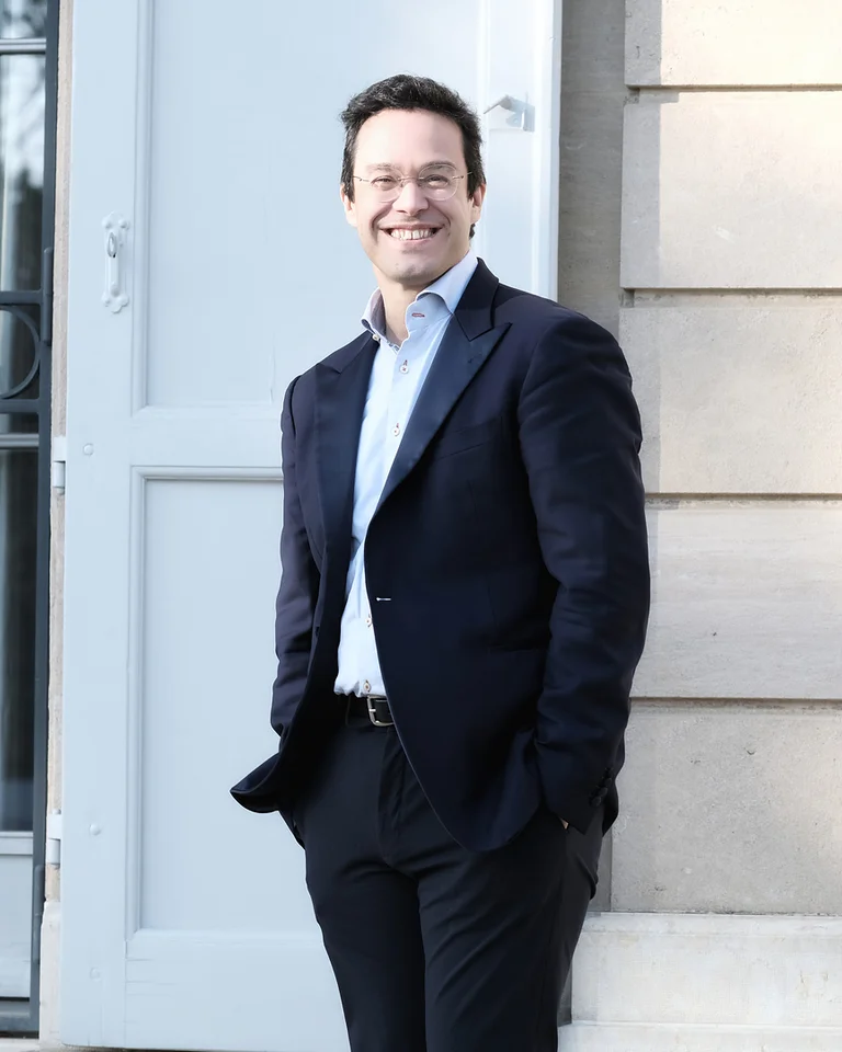 Ein Mann in einem dunklen Anzug (Leonardo García-Alarcón) steht lächelnd auf einer Straße vor einem Gebäude mit hellen Wänden und Tür.