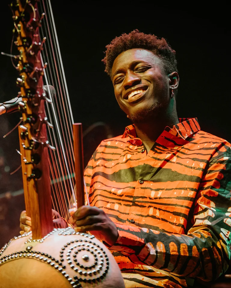 Momi Maiga spielt bei einem Auftritt auf einer Kora. Er trägt ein buntes Hemd und lächelt, während er spielt.