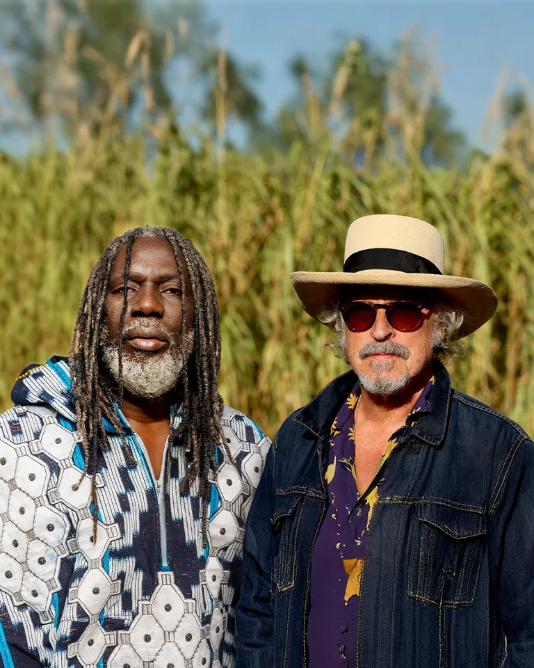 Zwei Männer (Tiken Jah Fakoly, Wolfgang Niedecken) stehen in einer sonnigen Landschaft mit hohem Gras und einem Fluss im Hintergrund, lässig gekleidet.