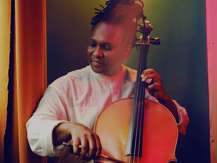 Ein Musiker (Abel Selaocoe) spielt leidenschaftlich Cello in einem warm beleuchteten Raum. Seine Kleidung ist kreativ und auffällig gemustert.