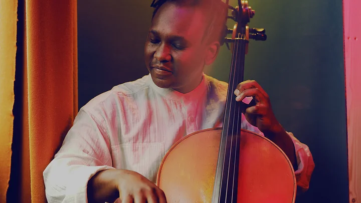 Ein Musiker (Abel Selaocoe) spielt leidenschaftlich Cello in einem warm beleuchteten Raum. Seine Kleidung ist kreativ und auffällig gemustert.