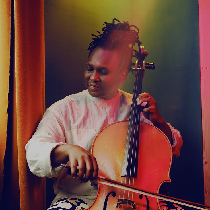 Ein Musiker (Abel Selaocoe) spielt leidenschaftlich Cello in einem warm beleuchteten Raum. Seine Kleidung ist kreativ und auffällig gemustert.