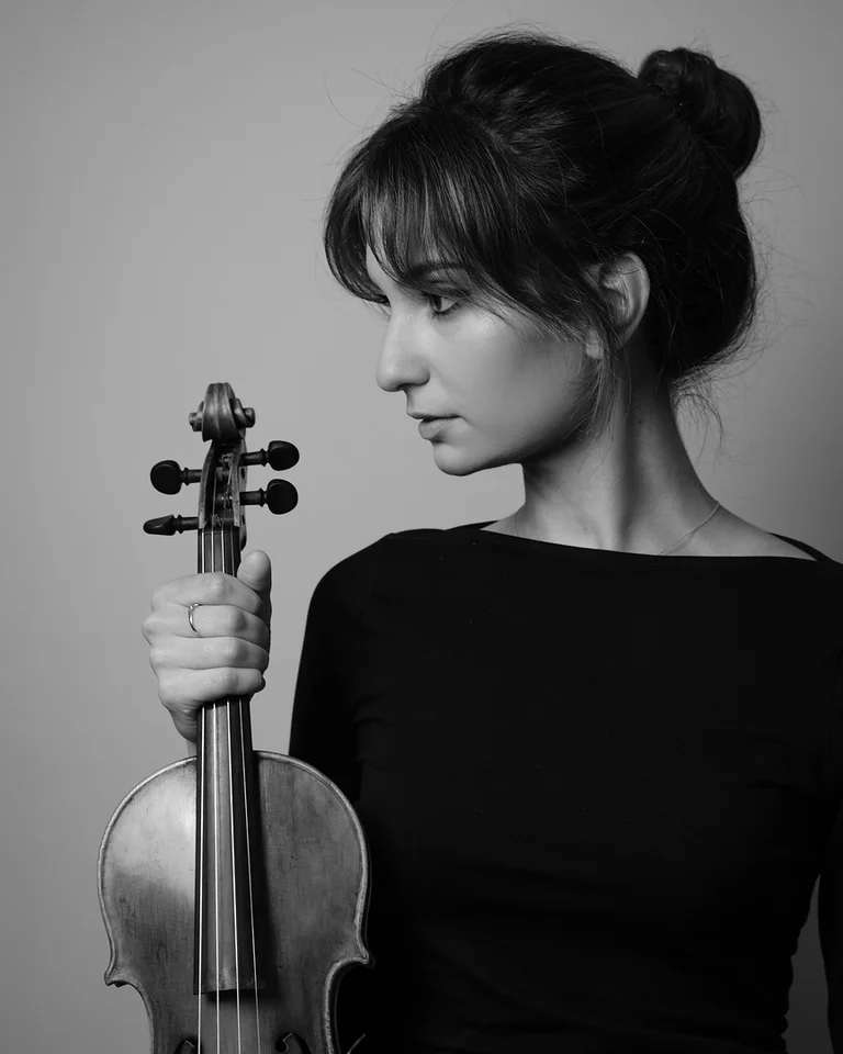 Schwarz-weiße Porträtaufnahme einer Frau (Ava Bahari) mit dunkel hochgestecktem Haar, die in ein langärmeliges Kleid gekleidet eine Geige hält.