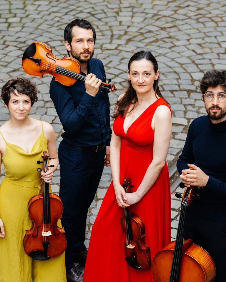 Vier Musiker stehen auf einem Kopfsteinpflaster. Zwei Männer und zwei Frauen halten Streichinstrumente und tragen elegante Kleidung.