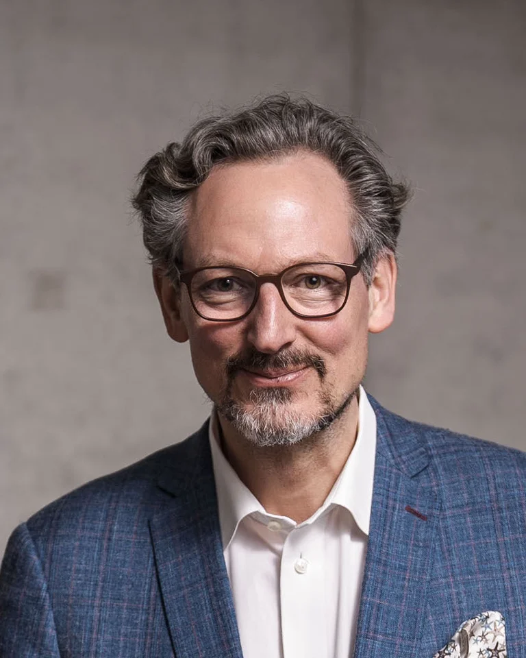 Ein Mann mittleren Alters mit Brille und lockigem Haar (Eckart von Hirschhausen) trägt einen blauen Anzug und ein weißes Hemd vor neutralem Hintergrund.
