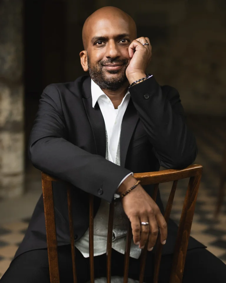 Ein Mann (Jonas Manuel Pinto) in einem Anzug sitzt auf einem Stuhl, seine Hand stützt sein Kinn. Der Hintergrund ist unscharf.