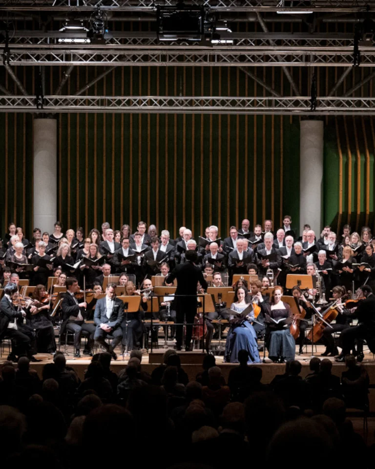 Ein großes Orchester spielt auf einer Bühne in einer Konzerthalle, mit vielen Musikern in formeller Kleidung, die verschiedene Instrumente spielen.