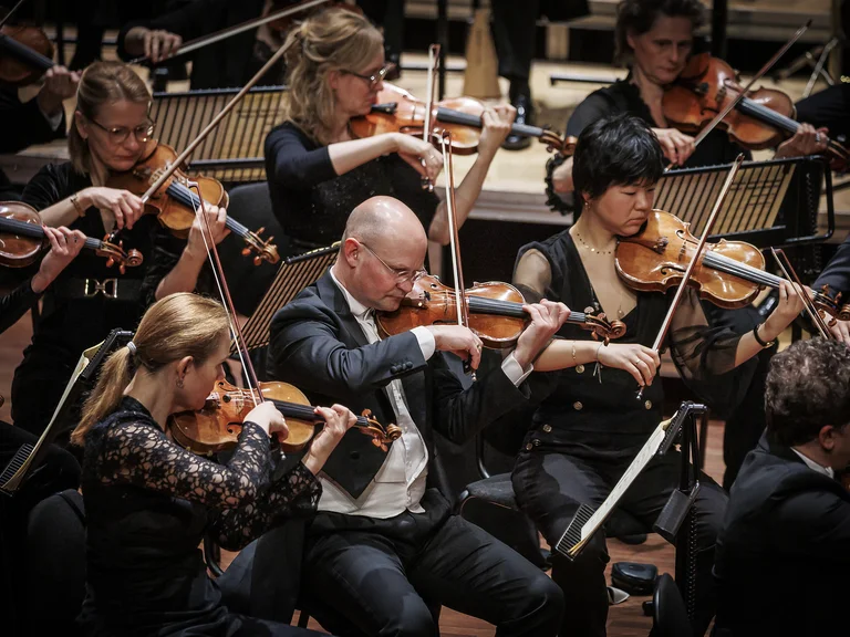 Ein Orchester voller Musiker spielt Geige auf der Bühne, alle in formeller Kleidung und konzentriert auf ihre Instrumente.