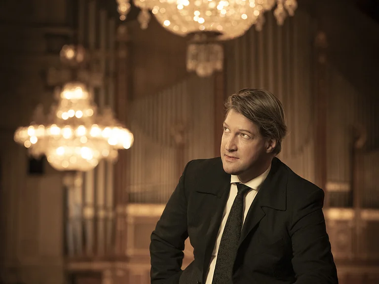 A stylishly dressed man (Gunther Rost) sits in an elegant hall with grand chandeliers, creating a noble and classical atmosphere.