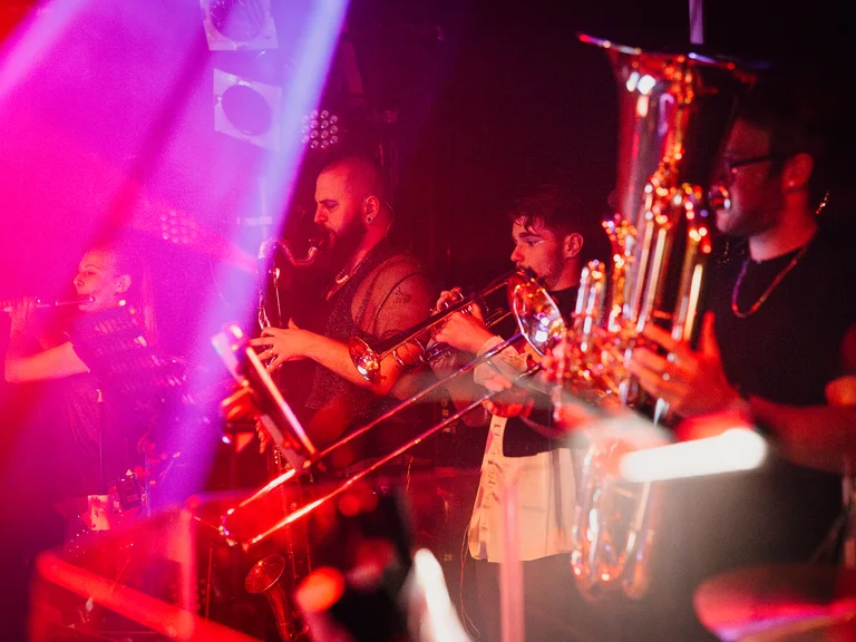 Eine Band (treppenhausorchester) spielt live auf der Bühne mit Saxophonen, umgeben von bunten Lichtern und lebendiger Atmosphäre.