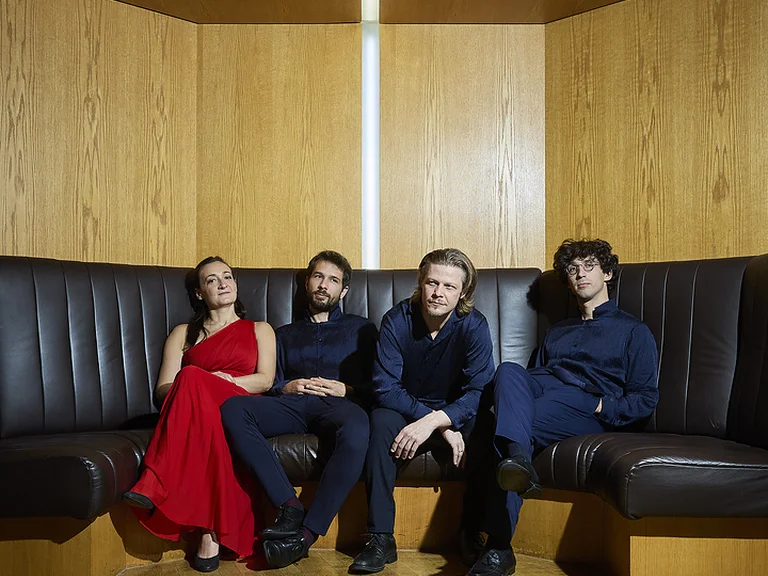Four people (Alinde Quartett) sit on the black leather sofa of the Kölner Philharmonie. A woman in a red dress is with three men in dark clothing.