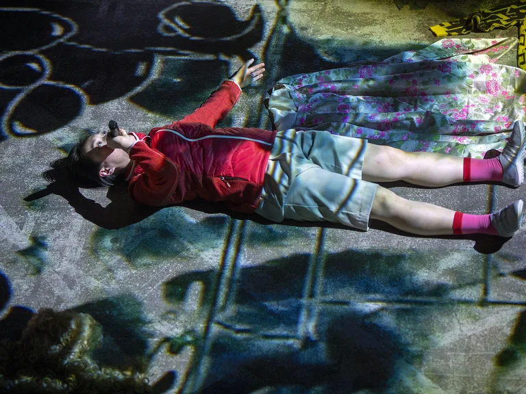 Person in bunter Projektion auf dem Boden liegend. Trägt rote und pinke Socken, Jeansshorts und sieht nachdenklich aus.