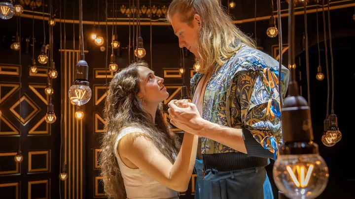 Zwei Personen in mittelalterlichen Kostümen halten sich an den Händen. Der Hintergrund ist mit warmen Lichtern geschmückt.