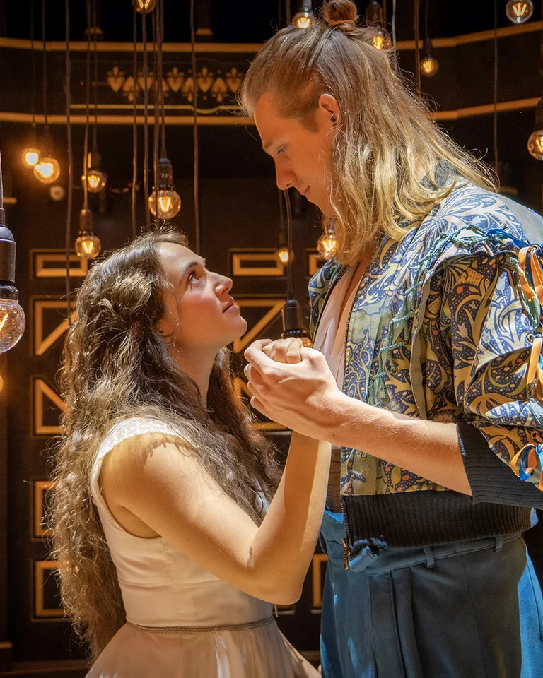 Zwei Personen in mittelalterlichen Kostümen halten sich an den Händen. Der Hintergrund ist mit warmen Lichtern geschmückt.