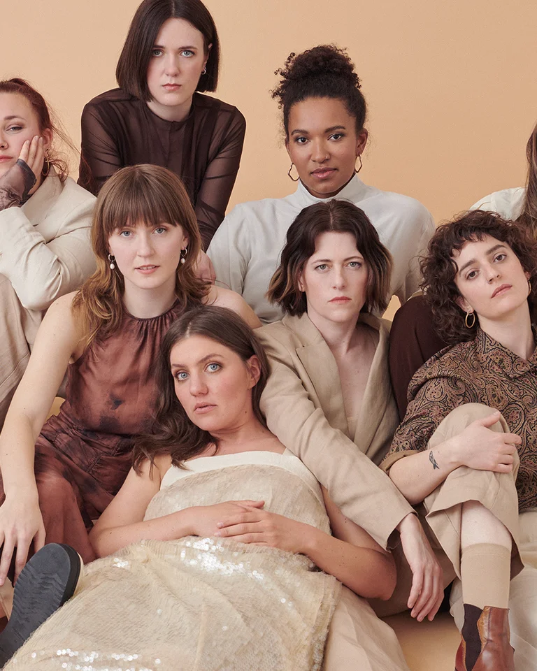 Eight people in neutral colors sit closely together against a beige background. They have relaxed, friendly expressions.
