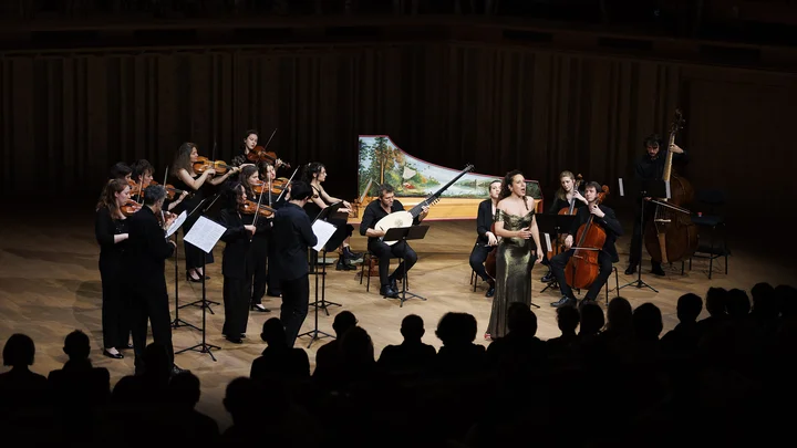 Ein Orchester mit Streichern und einem Cembalo spielt auf einer Bühne in einem dunklen Konzertsaal vor einem Publikum.