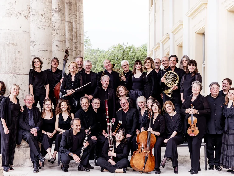 Eine große Gruppe von Musikern in Schwarz (Chamber Orchestra of Europe) steht vor einem historischen Gebäude. Verschiedene Instrumente wie Cello, Violine und Horn sind zu sehen.