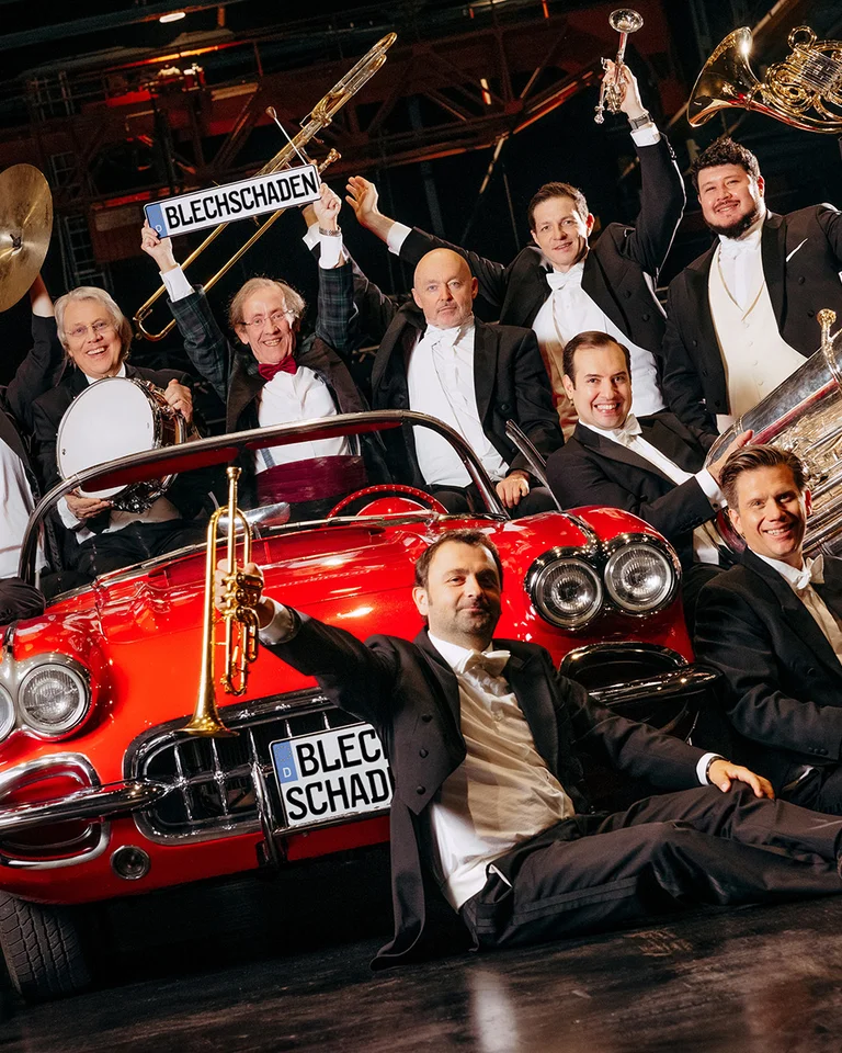 Eine Brass-Band posiert in eleganter Kleidung um ein rotes, klassisches Auto in einer Halle. Sie halten ihre Instrumente hoch.