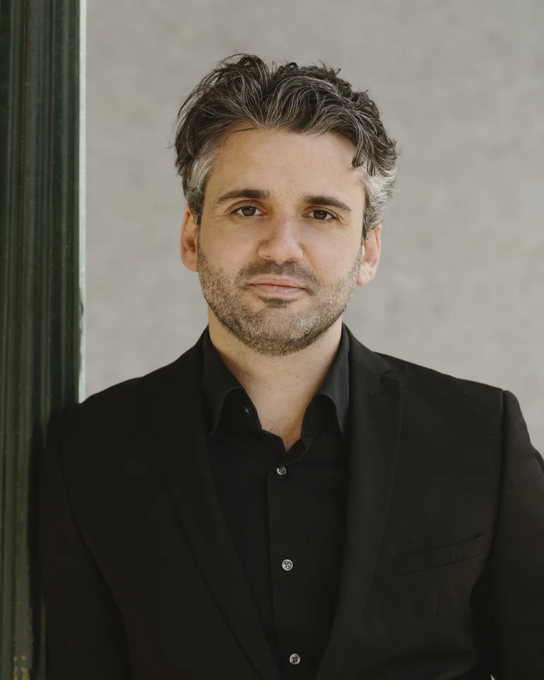 Ein Mann in einem schwarzen Anzug (Reinoud Van Mechelen) lehnt an einem Metallpfosten. Er hat graues Haar und einen ernsten Ausdruck.