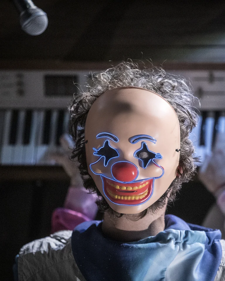 Eine Person mit Clownsmaske spielt auf einem Keyboard. Die Szene ist schummrig beleuchtet und wirkt geheimnisvoll.