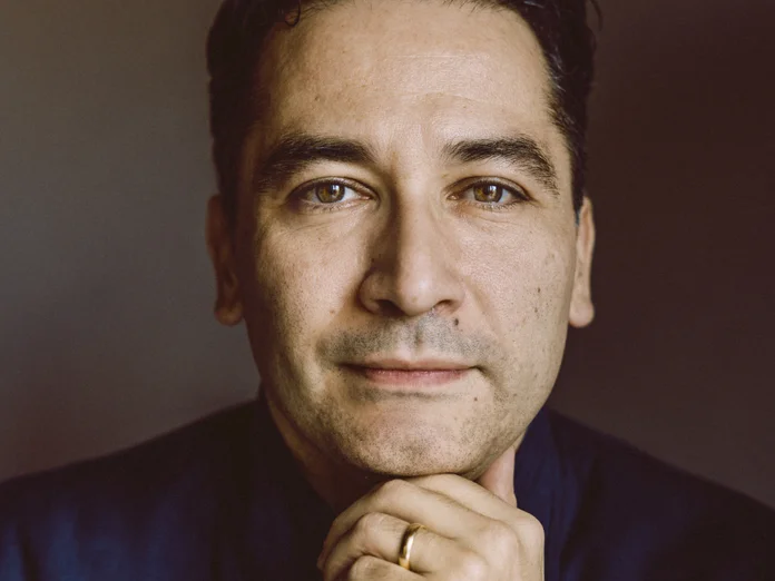 Porträt eines Mannes mit dunklem Haar, der nachdenklich in die Kamera schaut. Er trägt einen dunklen Anzug.