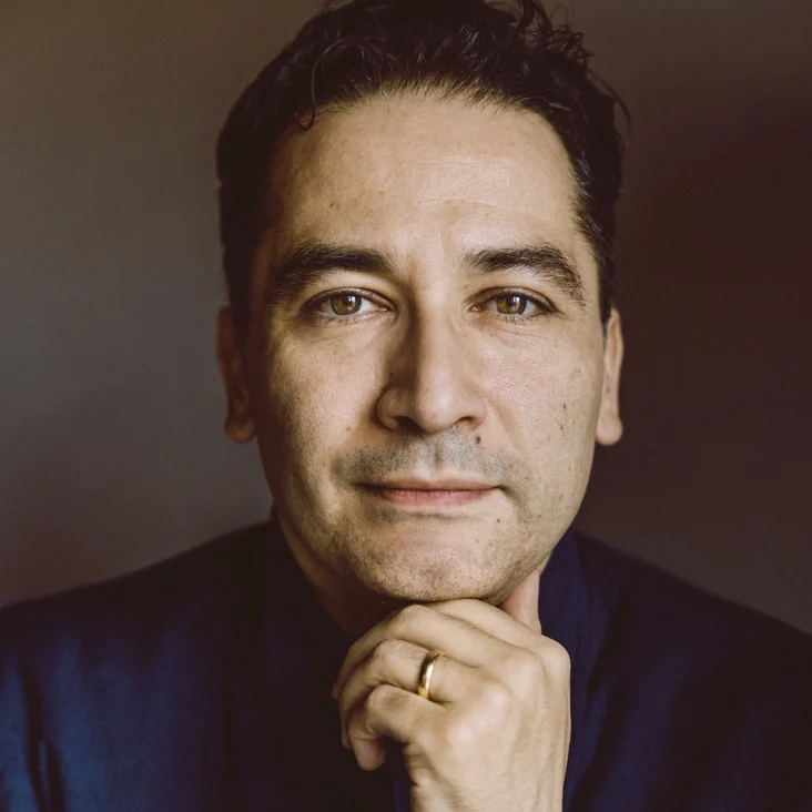 Porträt eines Mannes mit dunklem Haar, der nachdenklich in die Kamera schaut. Er trägt einen dunklen Anzug.