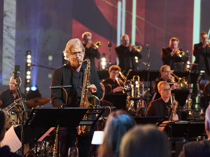 Ein Saxophonist spielt in einem Orchester mit Bläsern und Percussionisten (WDR Big Band). Die Bühne ist in stimmungsvolles Licht getaucht.