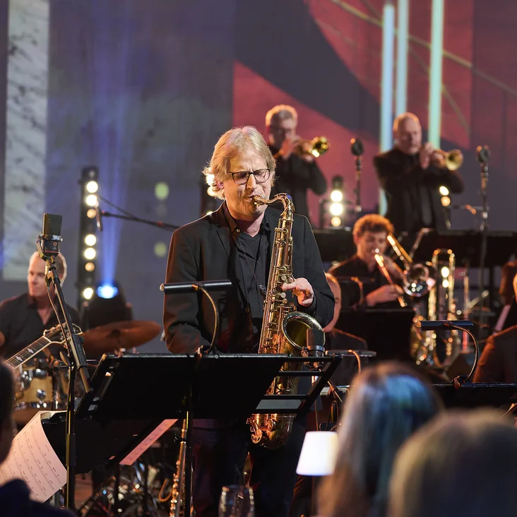 Ein Saxophonist spielt in einem Orchester mit Bläsern und Percussionisten (WDR Big Band). Die Bühne ist in stimmungsvolles Licht getaucht.