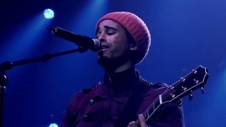 Patrice wearing a red beanie sings into a microphone on stage, accompanied by a guitar. Blue stage lights.