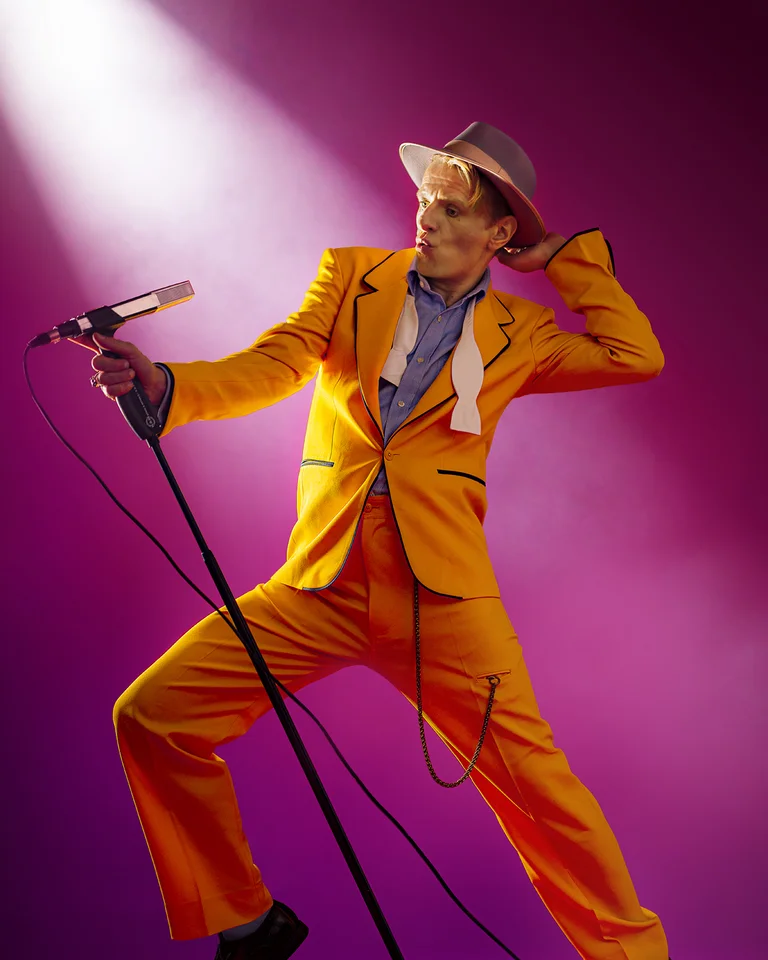 Alexander Scheer in einem auffälligen gelben Anzug und Hut, posiert dramatisch mit Mikrofon auf lilafarbenem Hintergrund. Show-Performance-Stimmung.