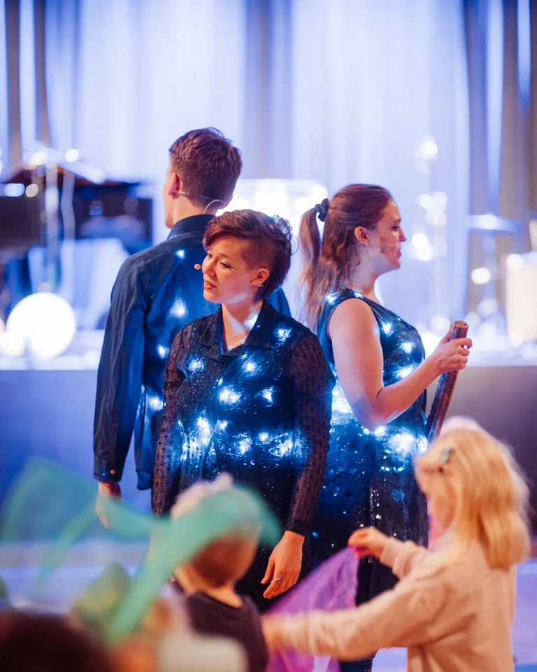 Künstler (Ensemble #kreuzvier ) tragen leuchtende Kostüme bei einer Bühnenshow. Im Vordergrund spielen Kinder mit bunten Tüchern. Die Atmosphäre ist lebhaft.