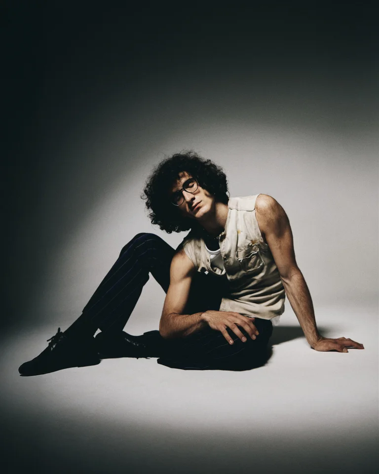 Eine Person mit lockigem Haar (Giorgi Gigashvili) sitzt lässig am Boden, elegant gekleidet in ein ärmelloses Oberteil und schwarze Hosen.