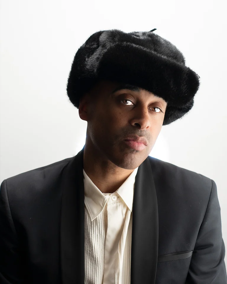 Mann (Patrice) in formellem Anzug und Pelzmütze, vor weißem Hintergrund. Seine Haltung ist ruhig und ausdrucksstark.