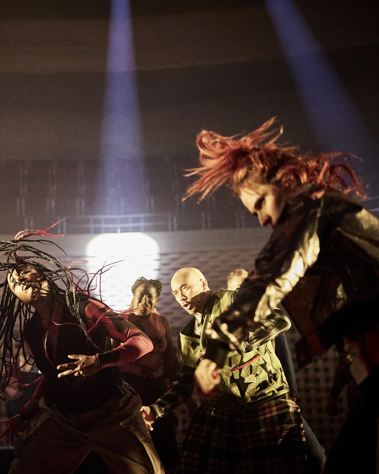 A group of dancers from the company Structure Rualitè in a dark hall, with dramatic lighting from above. The movement is intense and dynamic.